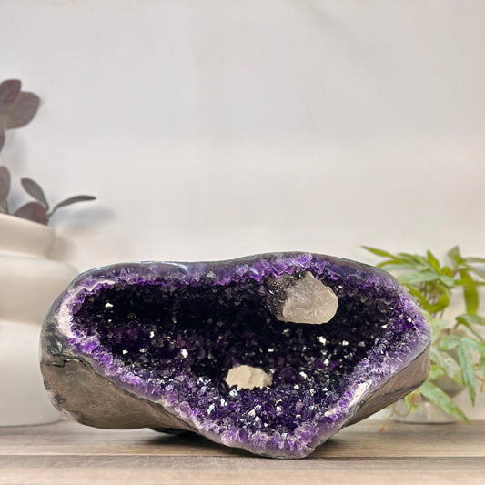 Weit geöffnete XXL Amethyst Geode aus Uruguay mit tiefvioletten Kristallspitzen und zwei prägnanten, hellen Kalzit-Formationen im Inneren auf einem Holzuntergrund.
